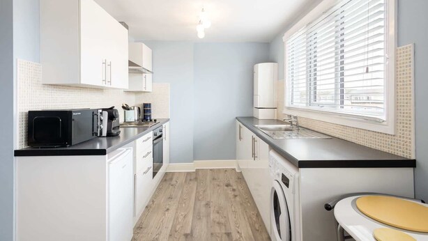 A compact kitchen with white cabinets, a microwave, a coffee maker, a washing machine, and a window with blinds.