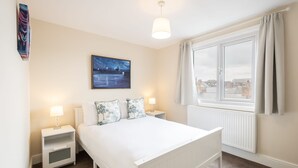 A bedroom with a bed, two bedside tables, a nightstand, a window with curtains, a radiator, and a painting on the wall.