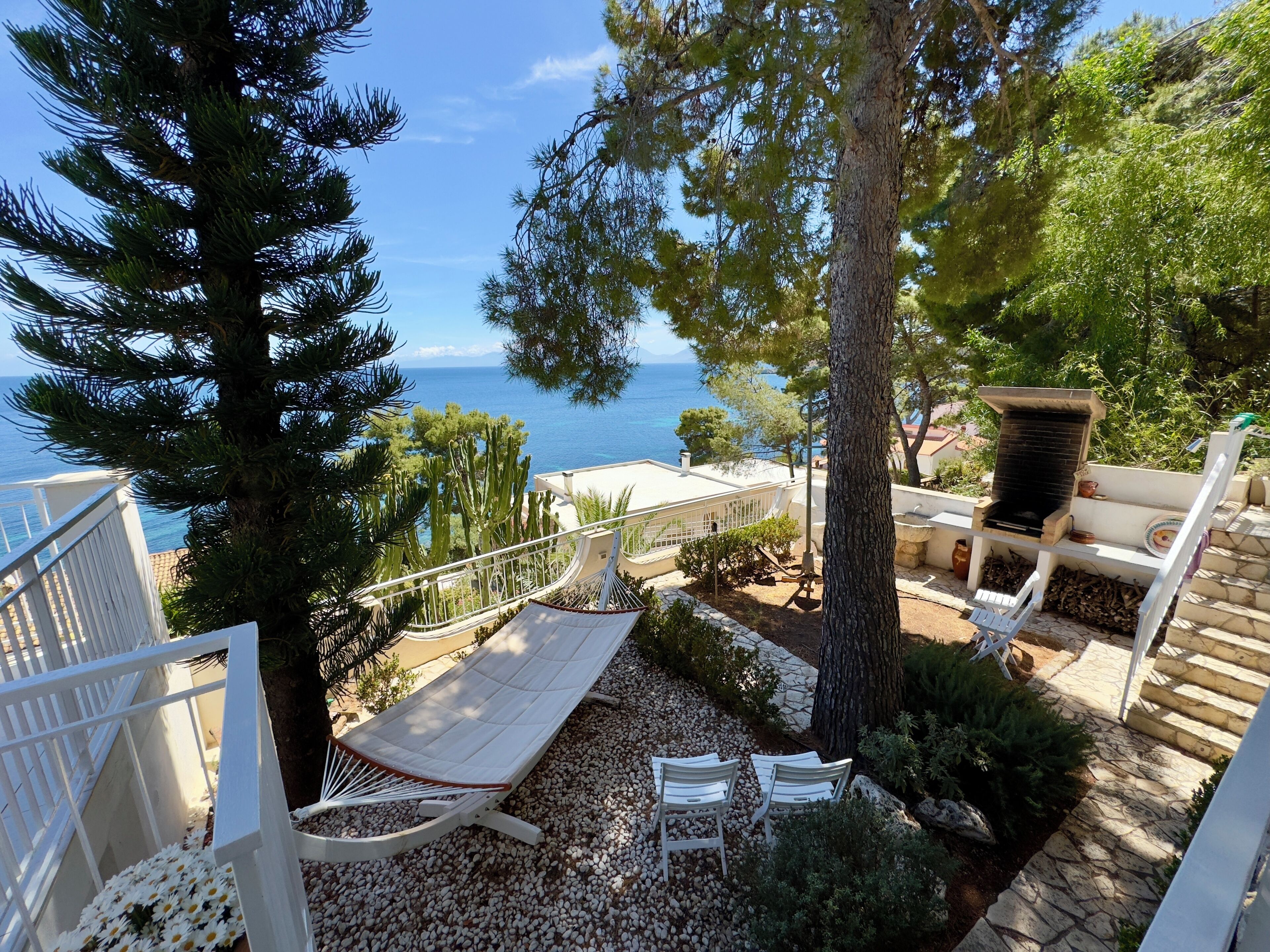 Uma rede, cadeiras brancas e um churrasqueira num jardim com vista para o mar.