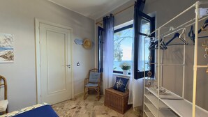 Een kamer met een bed, een stoel, een raam met gordijnen, een plank met spullen eraan en een deur.