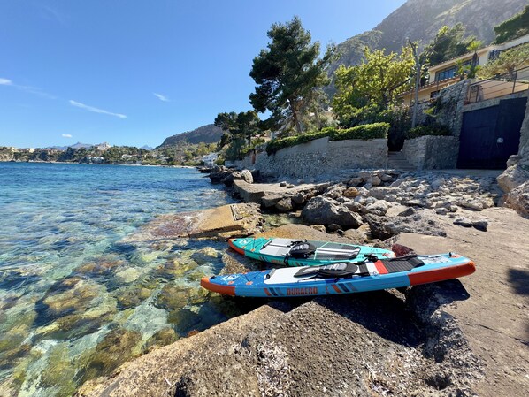 Orilla rocosa con agua cristalina, dos kayaks y un edificio con pared de piedra y vegetación.