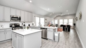 A modern kitchen with white cabinetry, a central island, stainless steel appliances, and a dining area with a black sofa and table. - Exquisite 4 Bedroom Brand New Home in Katy (Katy)