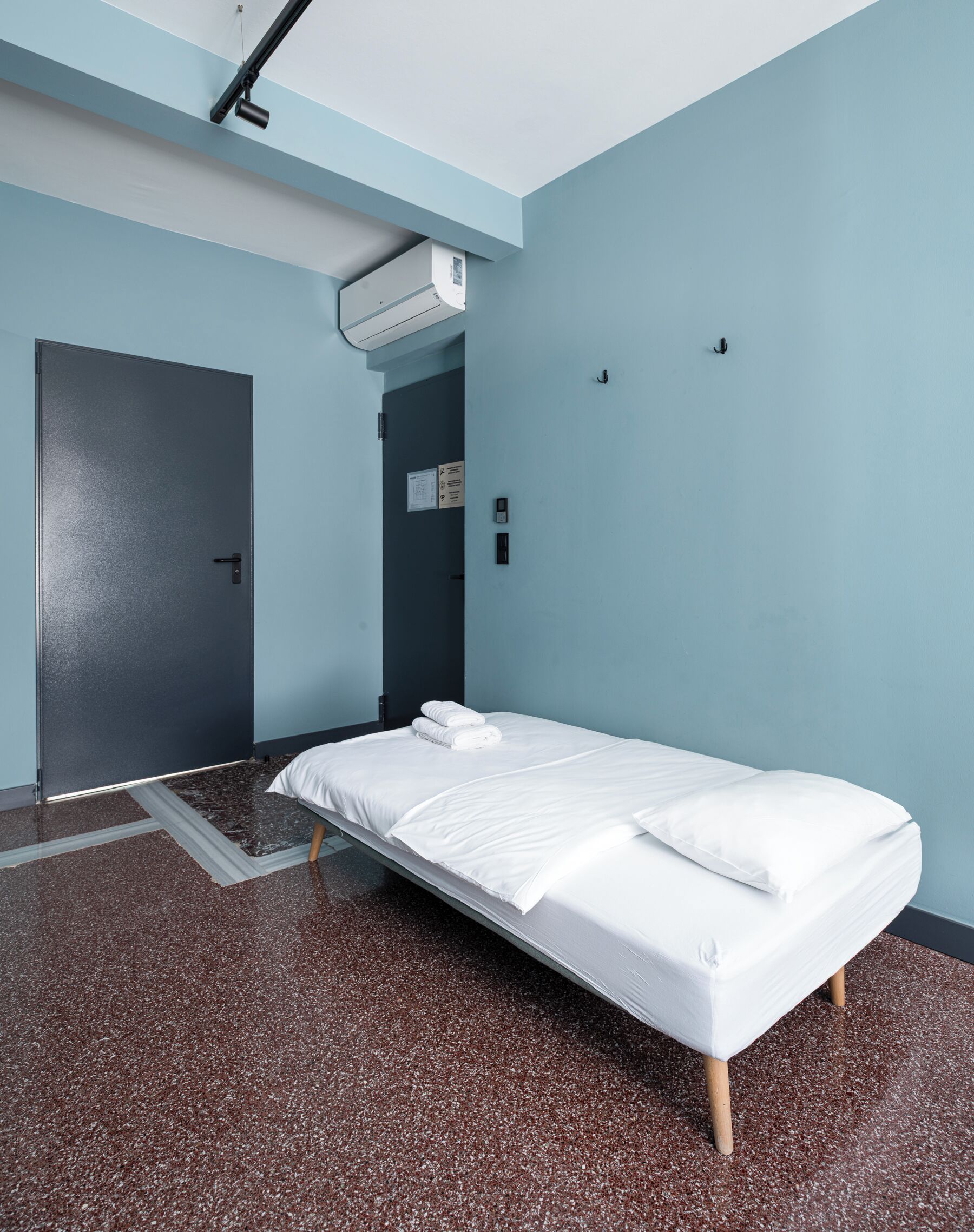 Ein Einzelbett in einem Zimmer mit dunklem Boden, hellblauen Wänden und einer Decken-Klimaanlage.