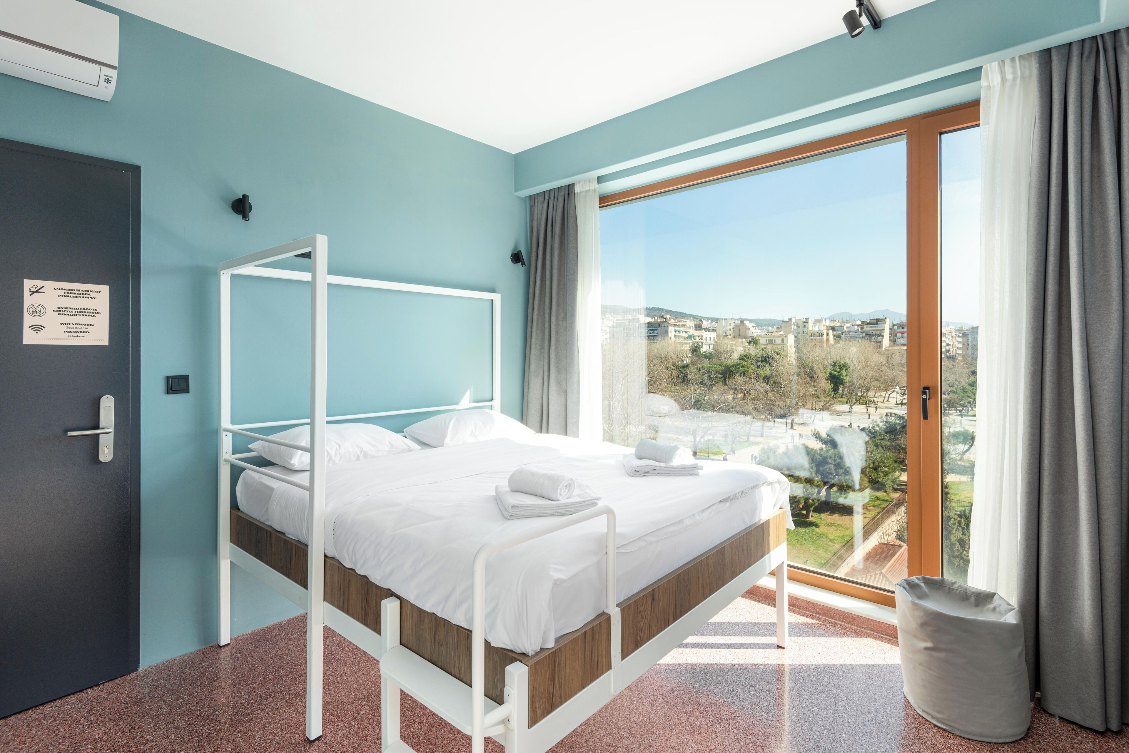 A modern hotel room with a bed, a view of the city, and a door with a sign.