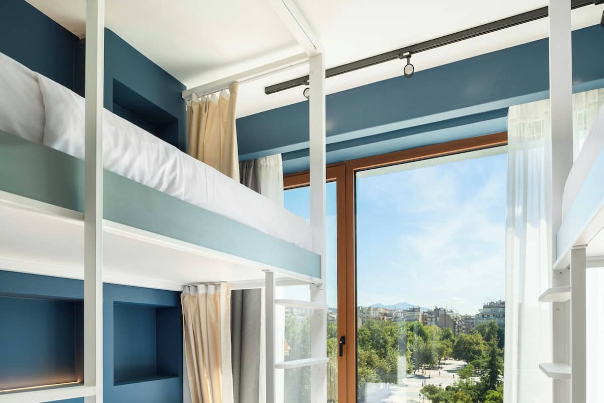 A room with bunk beds, a window with a city view, and a small trash bin.