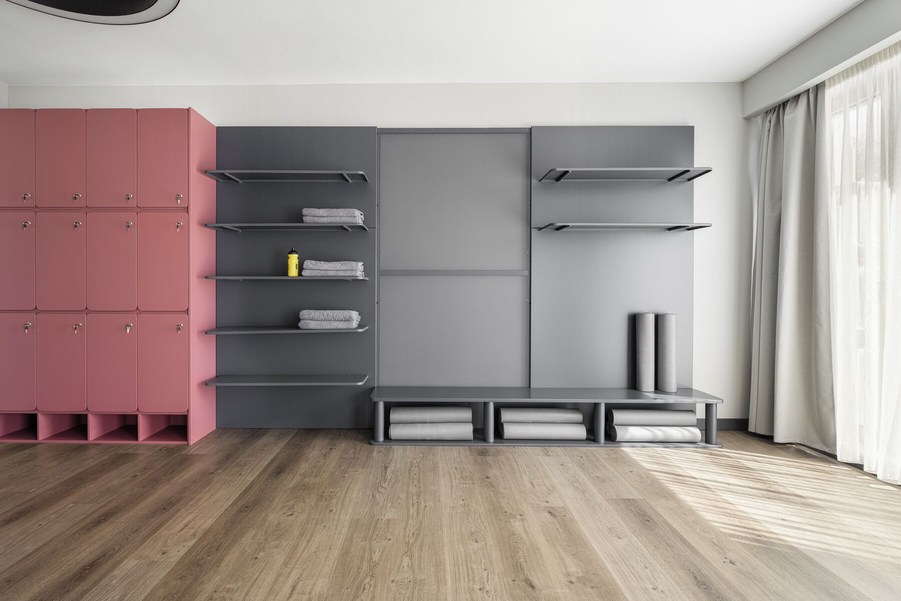 A modern room with a grey wardrobe, pink storage units, and a grey shelving unit with towels and a water bottle.