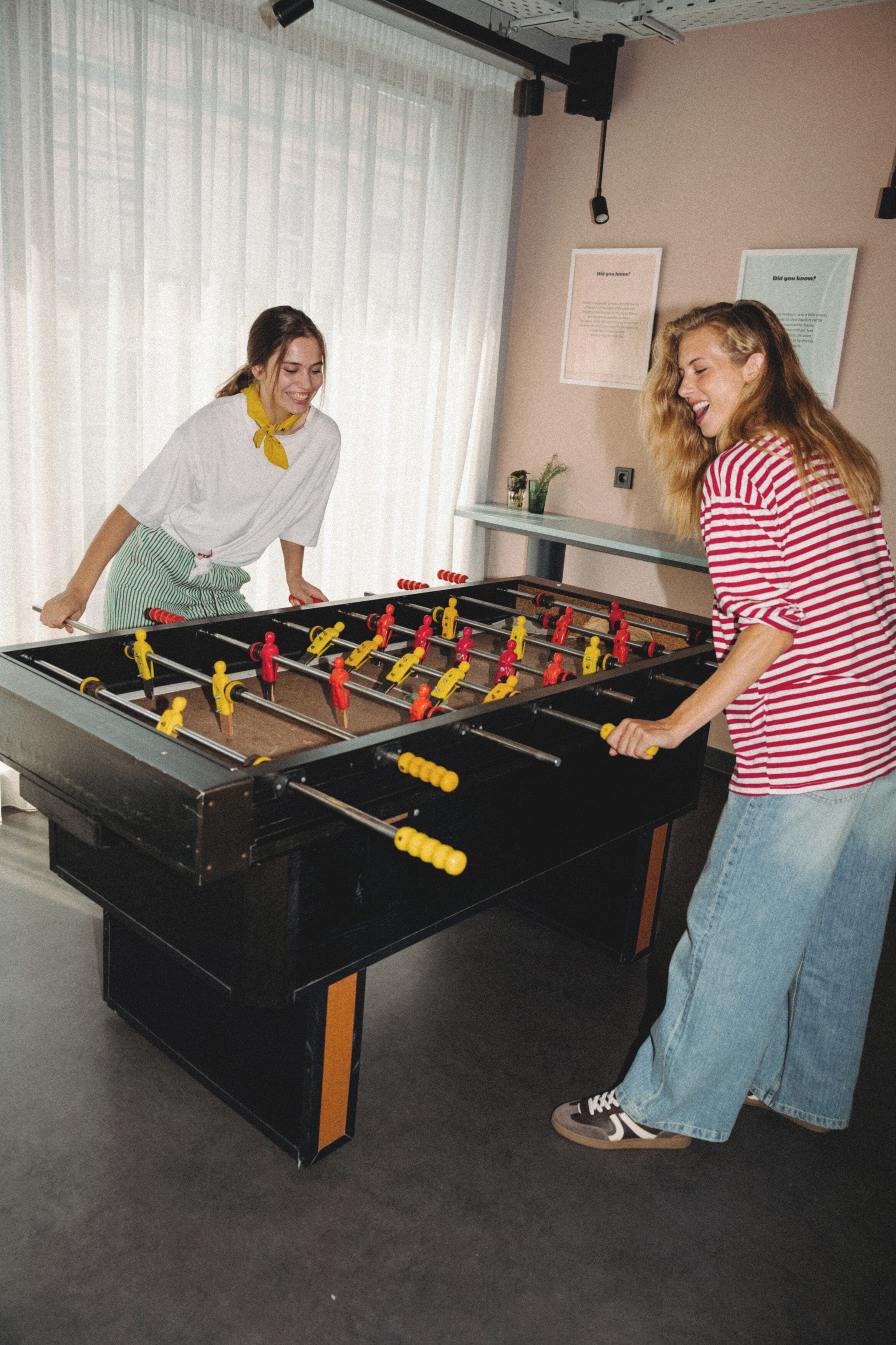Two people playing foosball in a room with a large table, a window with curtains, and a wall with framed text.