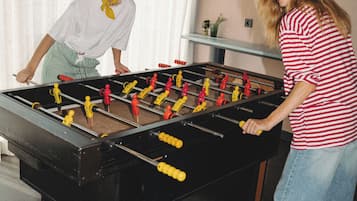 Two people playing foosball in a room with a large table, a window with curtains, and a wall with framed text.