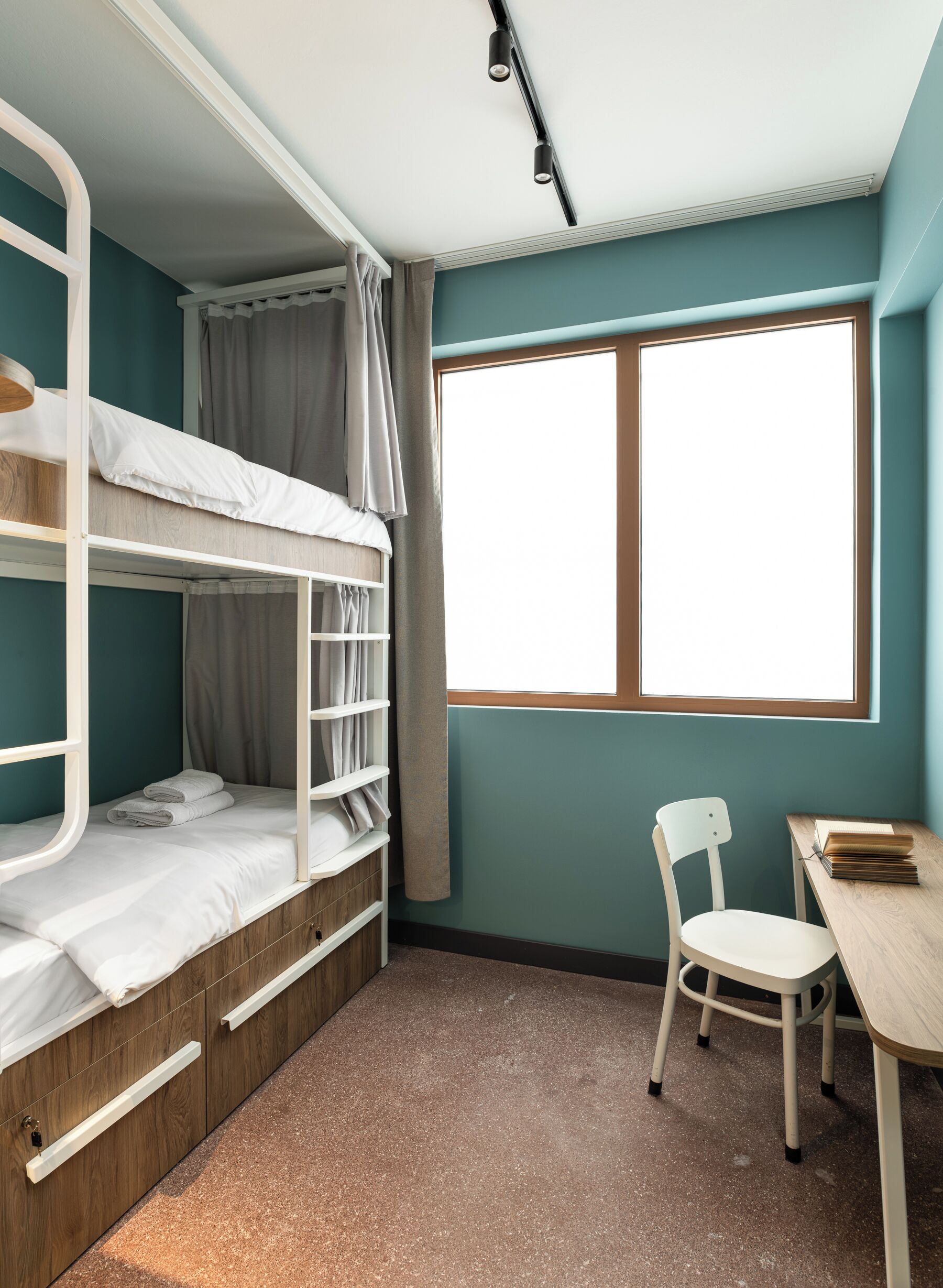A bunk bed room with a desk and chair, a window with curtains, and a wooden floor.