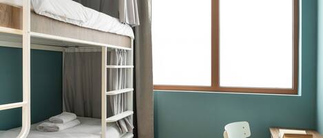 A bunk bed room with a desk and chair, a window with curtains, and a wooden floor.