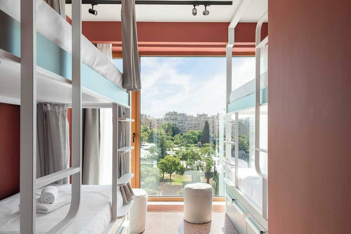 A modern hotel room with bunk beds, a glass partition, and a view of a cityscape.