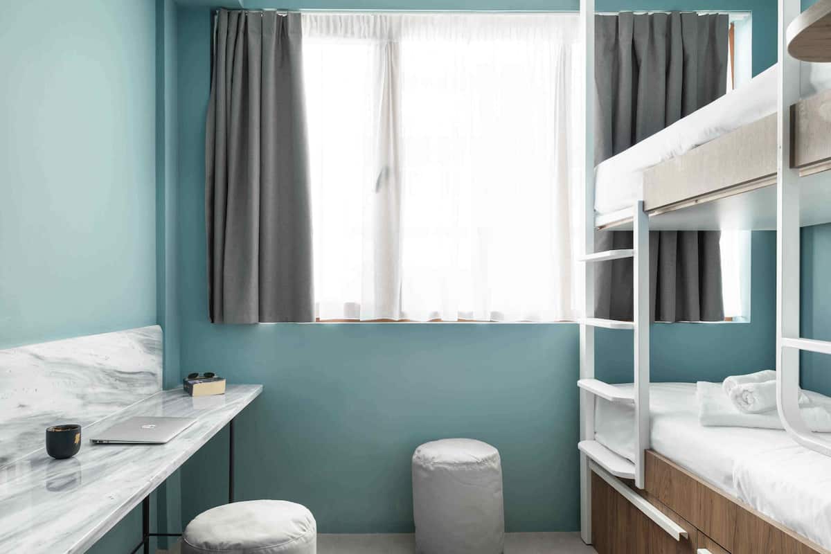A compact room with a bunk bed, a desk with a laptop, a small ottoman, and a window with curtains.