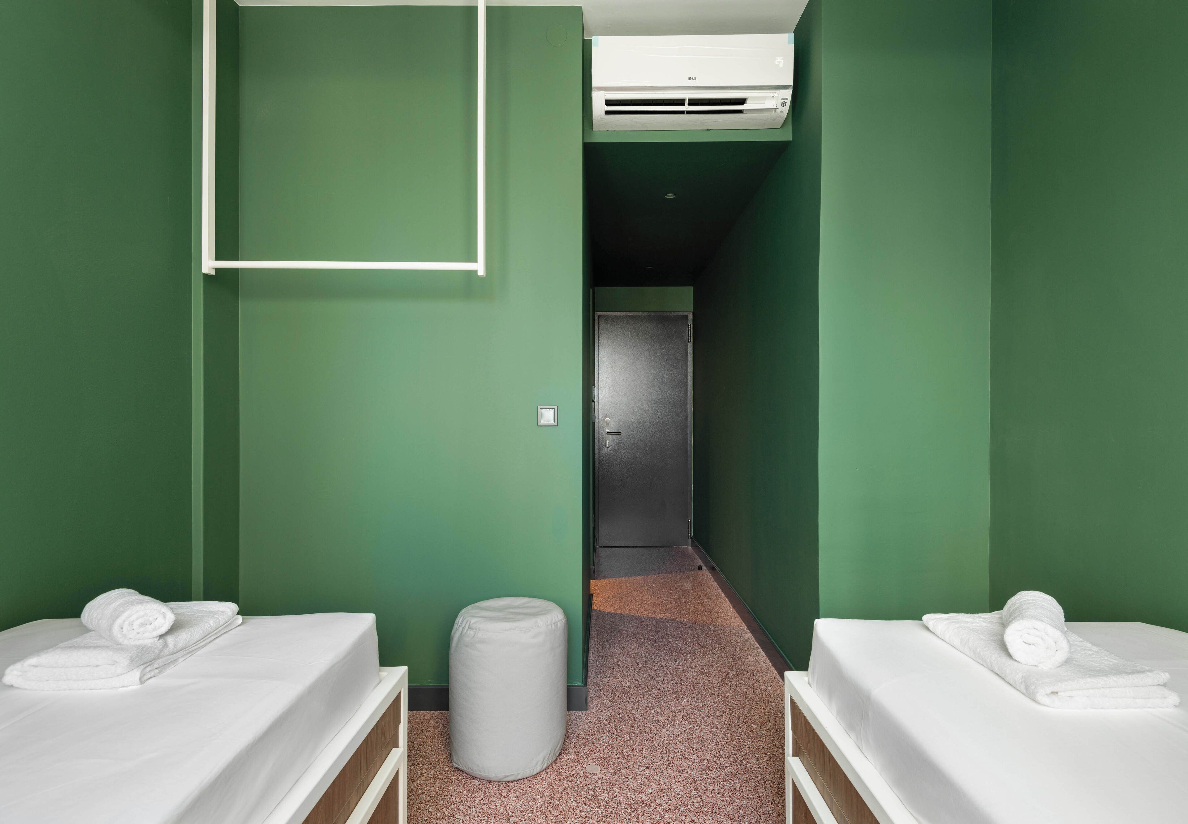 Una camera d'albergo con due letti, uno sgabello, un cestino e un condizionatore d'aria.