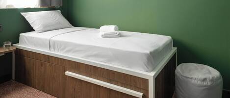 A single-bed room with a wooden frame, a white mattress, and a grey bean bag.