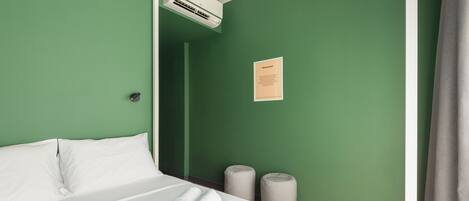 Habitación de hotel con pared verde, cama con ropa blanca, dos toallas enrolladas, unidad de aire acondicionado y un pequeño cartel en la pared.