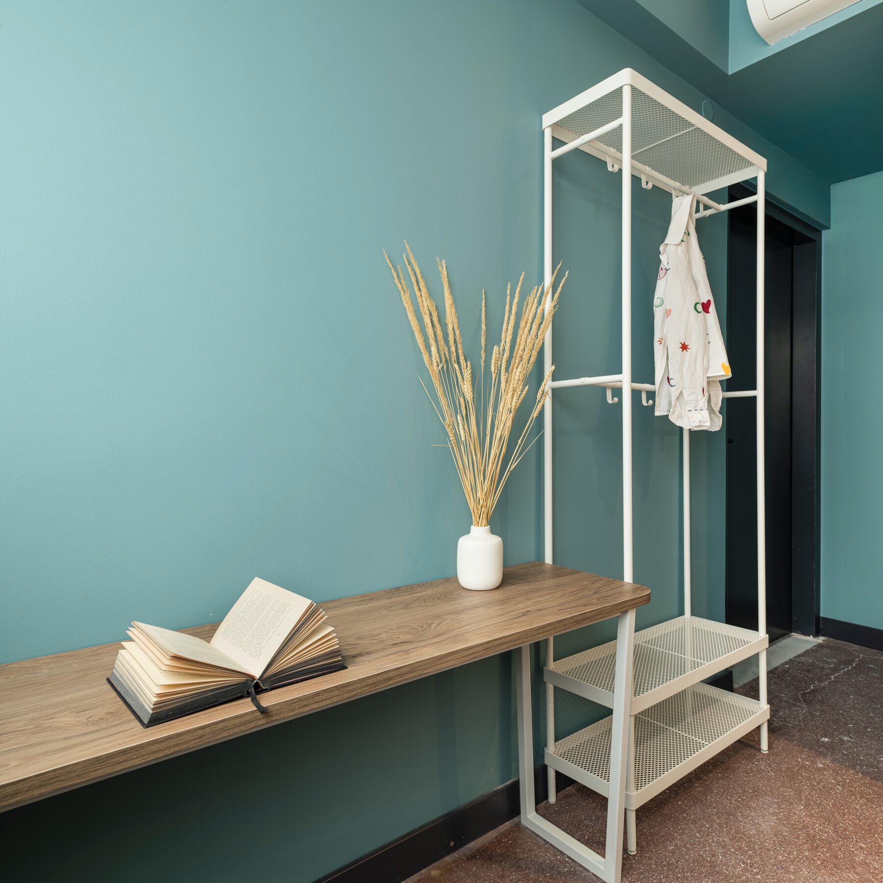 Una habitación con paredes color turquesa, un banco de madera, un jarrón blanco con plantas secas y una estantería metálica blanca con un abrigo colgado.