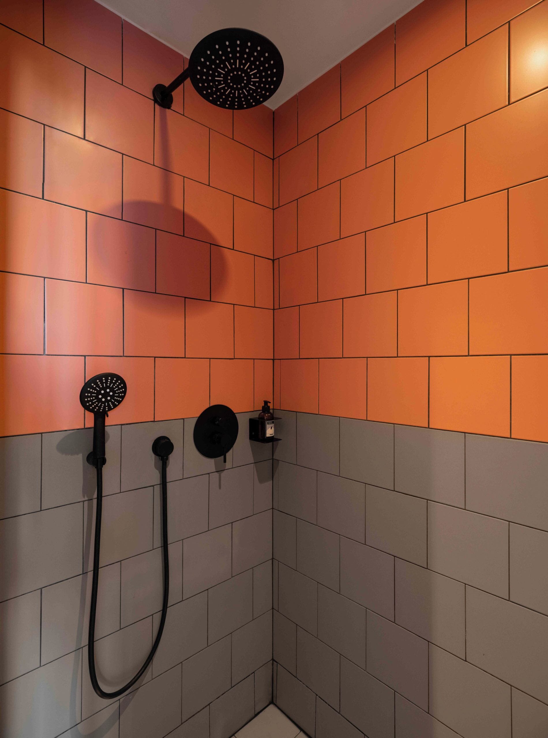 A shower with a black handheld showerhead and a wall-mounted head, both mounted on a tiled wall with orange and grey tiles.