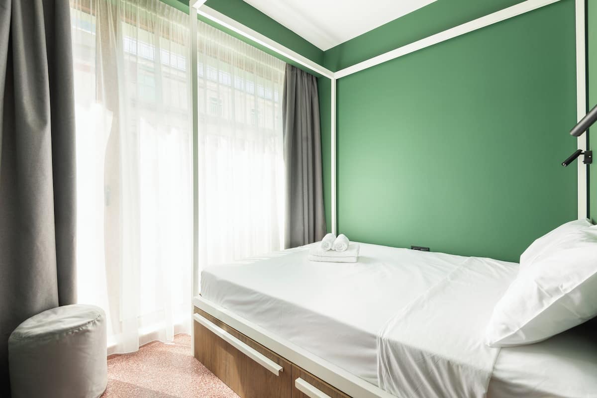 A neatly made bed with white linens, a small bedside table, and a window with sheer curtains.