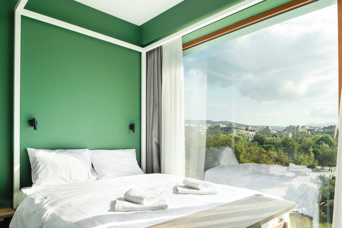 A modern bedroom with a bed, a wooden bedside table, and a large window offering a city view.