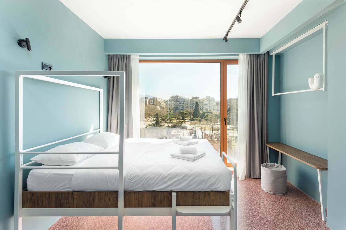 A modern bedroom with a bed, a window with a view of buildings, a bedside table, and a trash bin.