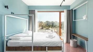 Un dormitorio moderno con cama, ventana con vista a edificios, mesita de noche y un cubo de basura.