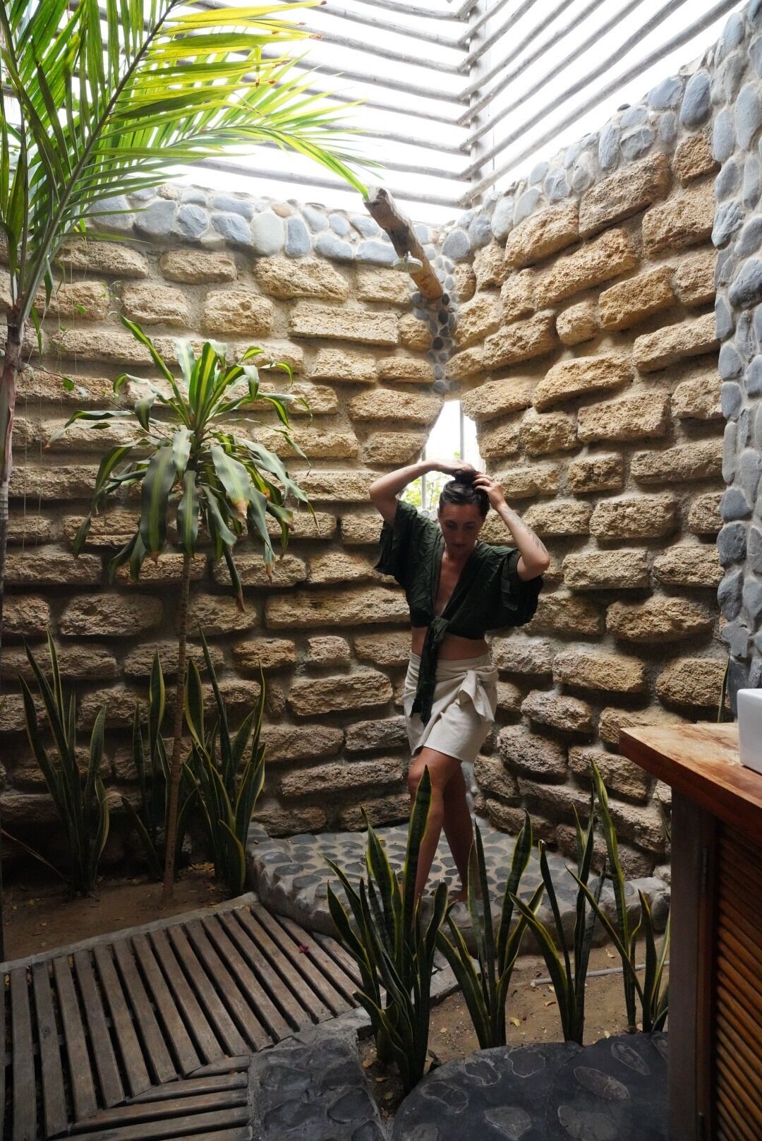 Una persona en una habitación de paredes de piedra y techo inclinado, rodeada de varias plantas.