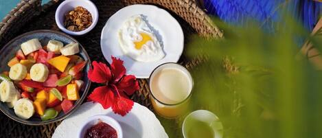 A person holding a wicker tray with breakfast items including fruit, eggs, bread, and beverages.
