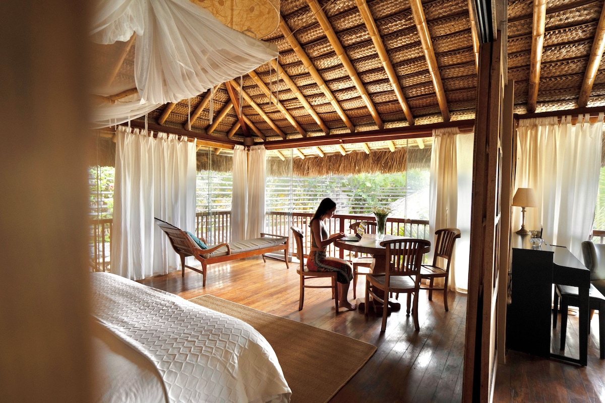 A room with a bed, a dining table, and chairs, with a view of greenery outside.