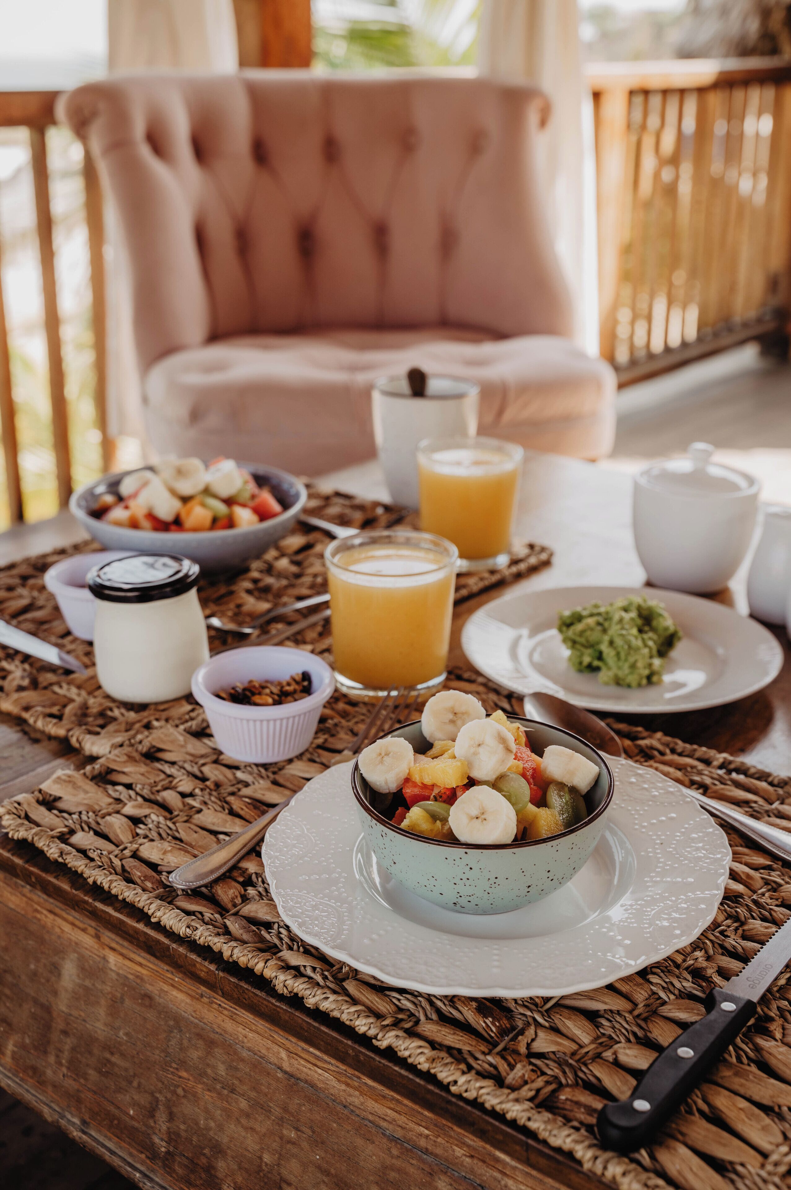 Un desayuno dispuesto sobre una mesa de madera con un mantel tejido, que incluye tazones de fruta, un vaso de jugo de naranja, una taza de café y una rebanada de pan tostado con aguacate.