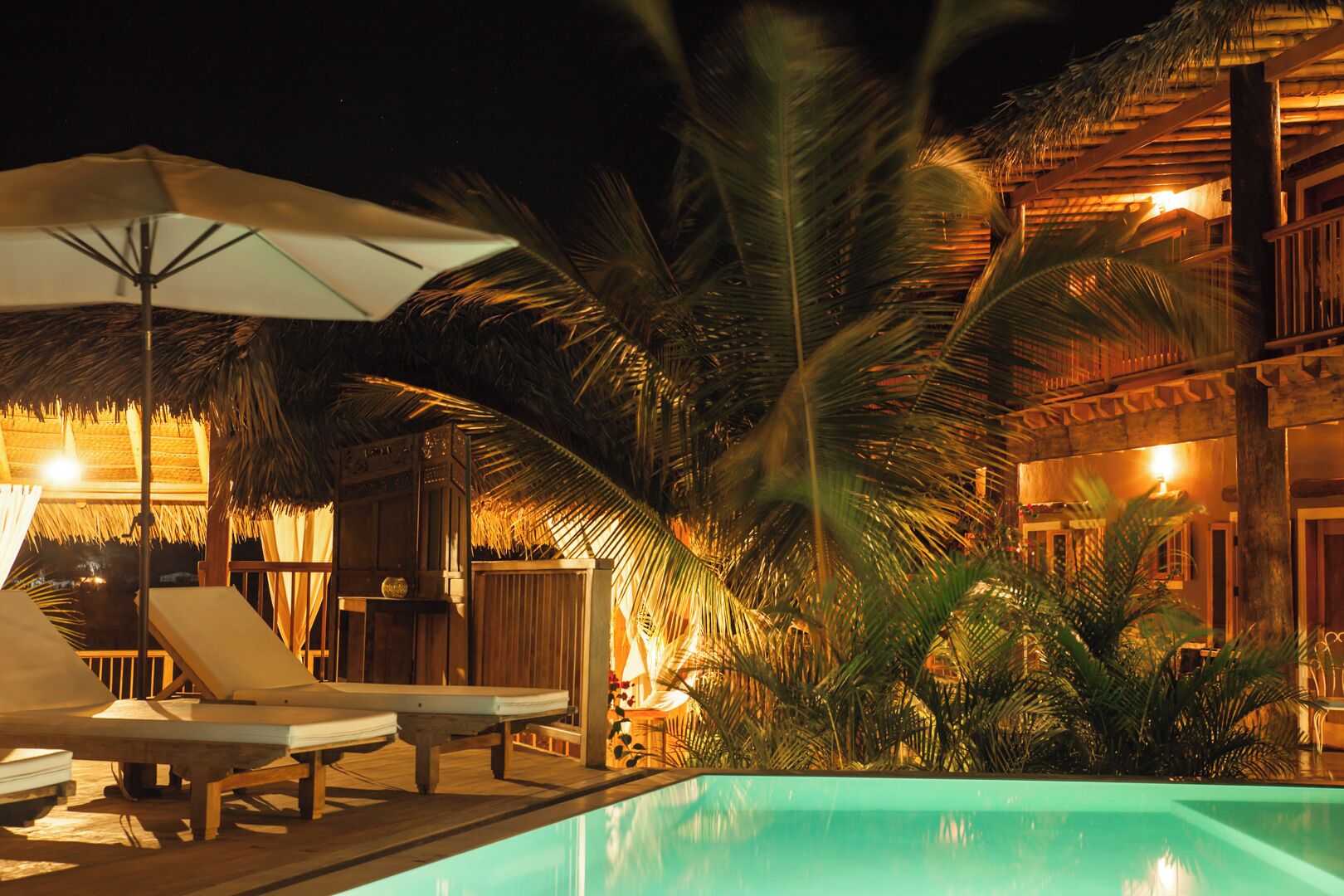 Zona de piscina con sillas de descanso y sombrillas, rodeada de palmeras y un edificio con iluminación cálida.