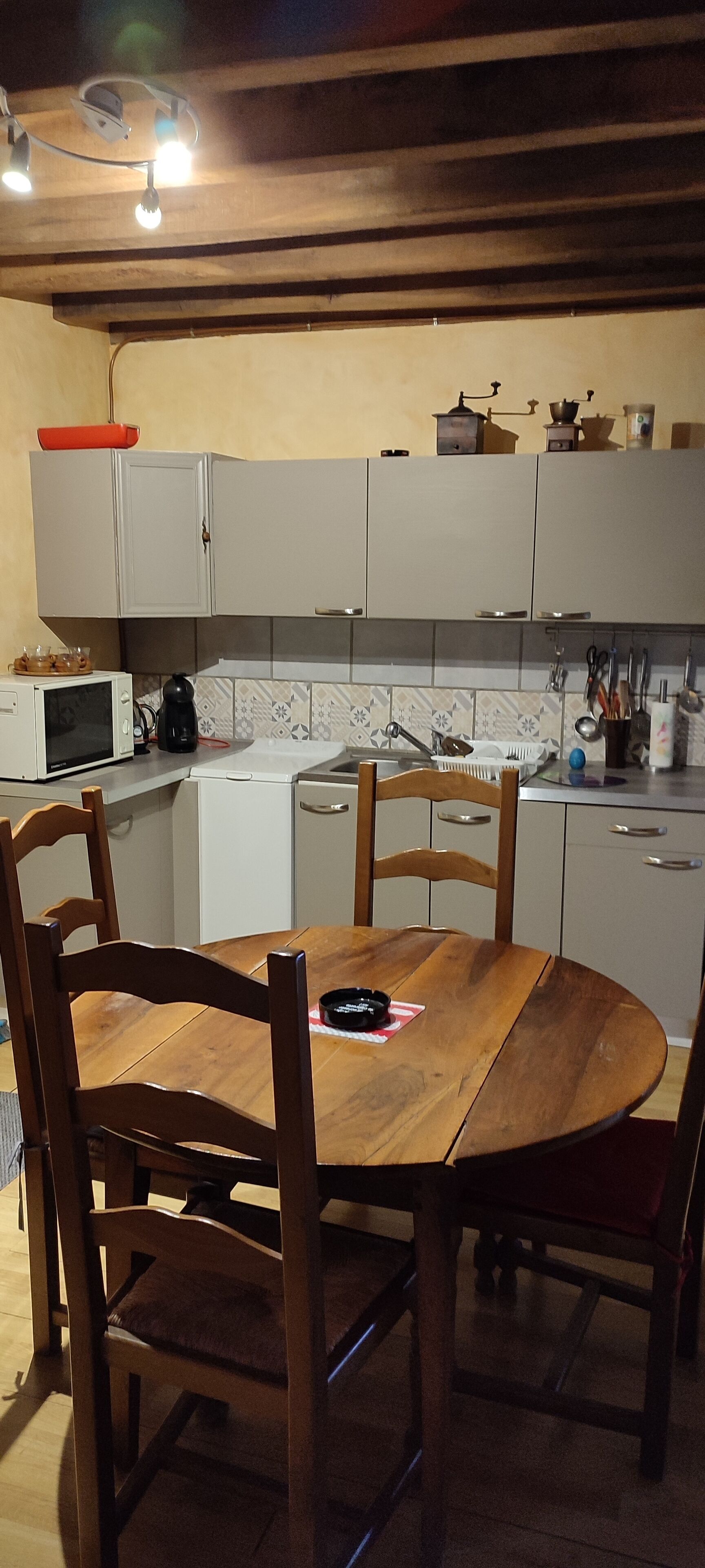 Eine Küche mit weißen Schränken, einem Holzespeise Tisch und Stühlen. Auf der Arbeitsplatte befinden sich eine Mikrowelle, ein Wasserkocher und verschiedene Küchenutensilien.