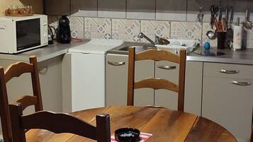 Eine Küche mit weißen Schränken, einem Holzespeise Tisch und Stühlen. Auf der Arbeitsplatte befinden sich eine Mikrowelle, ein Wasserkocher und verschiedene Küchenutensilien.