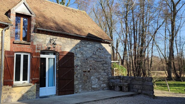 A stone building with a wooden door and window shutters, surrounded by trees.