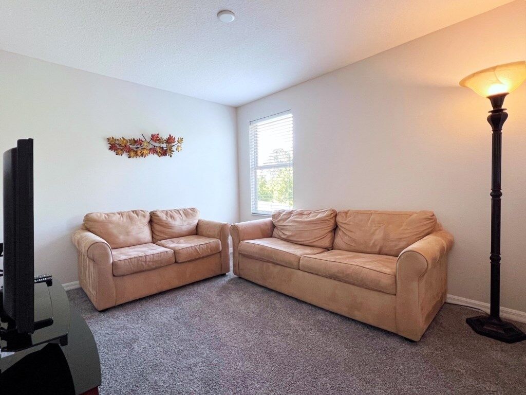 A living room with two beige leather sofas, a flat-screen TV, a decorative leaf wreath, a standing floor lamp, and a window with blinds.