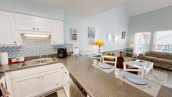 A modern kitchen with white cabinets, a granite countertop, and a coffee maker. There is a dining area with a patterned sofa and a lamp.