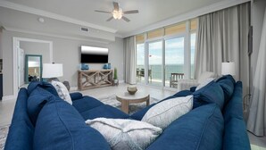 A spacious living room with a large blue sofa, a flat-screen TV, and a view of the sea through large windows.