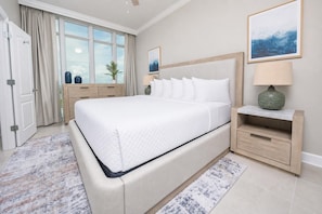 A bedroom with a large bed, a nightstand, a lamp, a window with curtains, and a door.