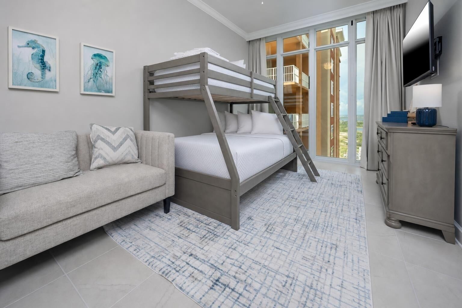 A modern hotel room with a bunk bed, a sofa, a flat-screen TV, and a large window with a view.