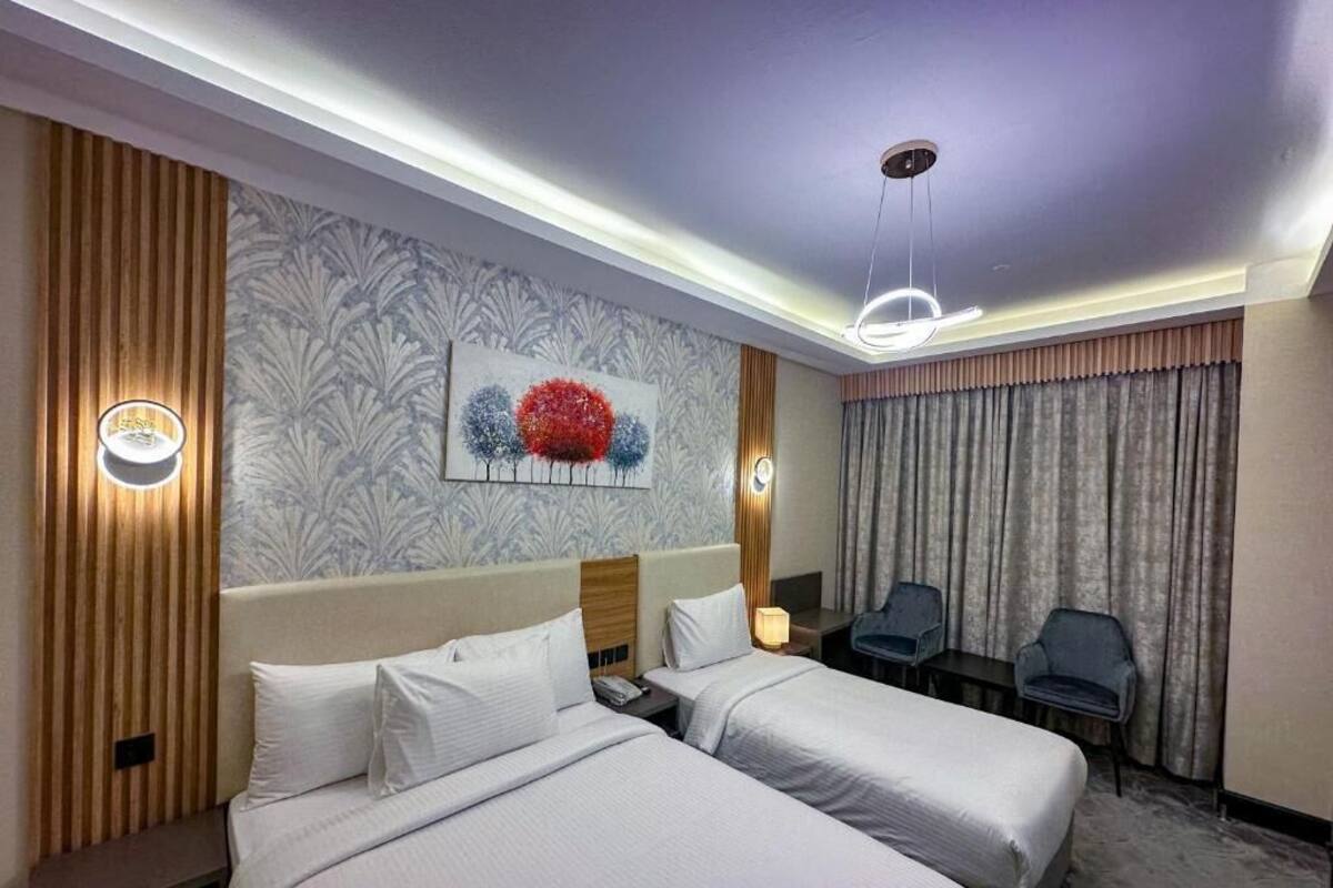A hotel room with two beds, a headboard, a wall-mounted artwork, a ceiling light, a nightstand, a chair, and a window with curtains.