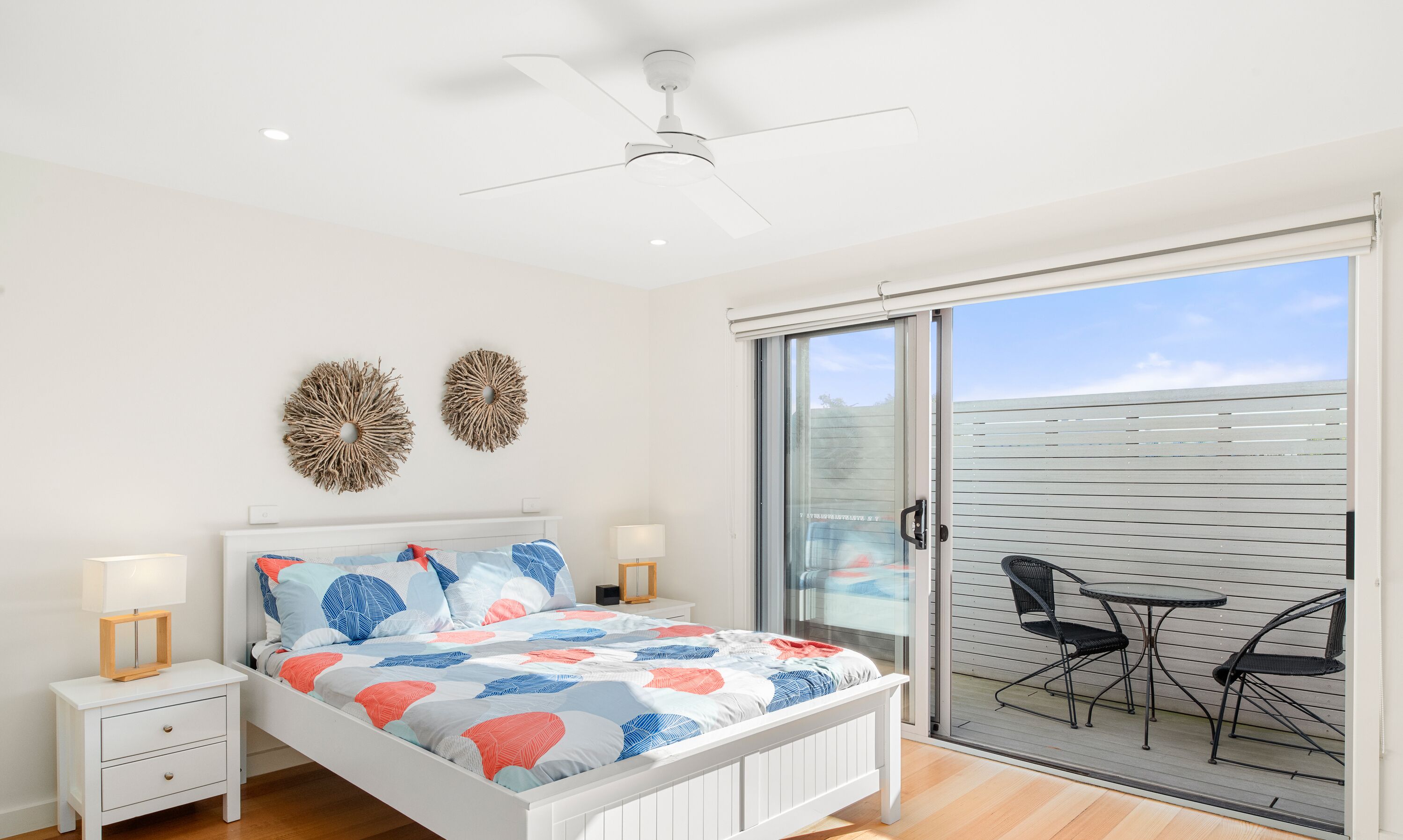 Un dormitorio con cama, mesitas de noche, ventilador de techo y una puerta corrediza de vidrio que da a un espacio exterior con sillas.