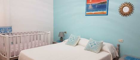 A bedroom with a white crib, a bed with a white bedspread, two decorative pillows, a nightstand with a lamp, and a wall-mounted sun decoration.