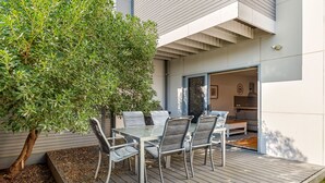 Un patio moderno al aire libre con una mesa de vidrio y sillas de metal, rodeado por una terraza de madera y un árbol grande.