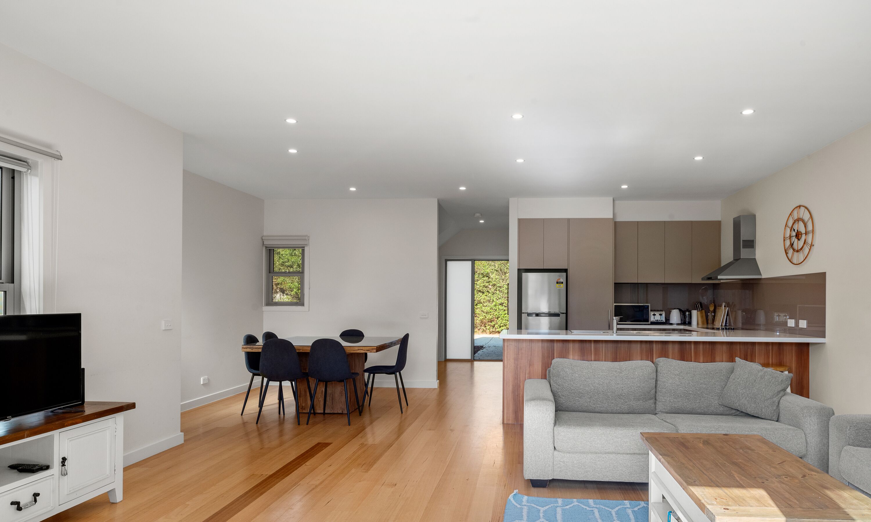 A modern living room with a grey sofa, wooden coffee table, and a TV on a wooden stand. There is a dining area with a table and chairs, and a kitchen with a microwave and stove.