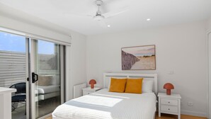 Une chambre à coucher comprenant un lit, une table de chevet, une lampe, une porte-fenêtre coulissante et un tableau représentant une scène de plage au mur.