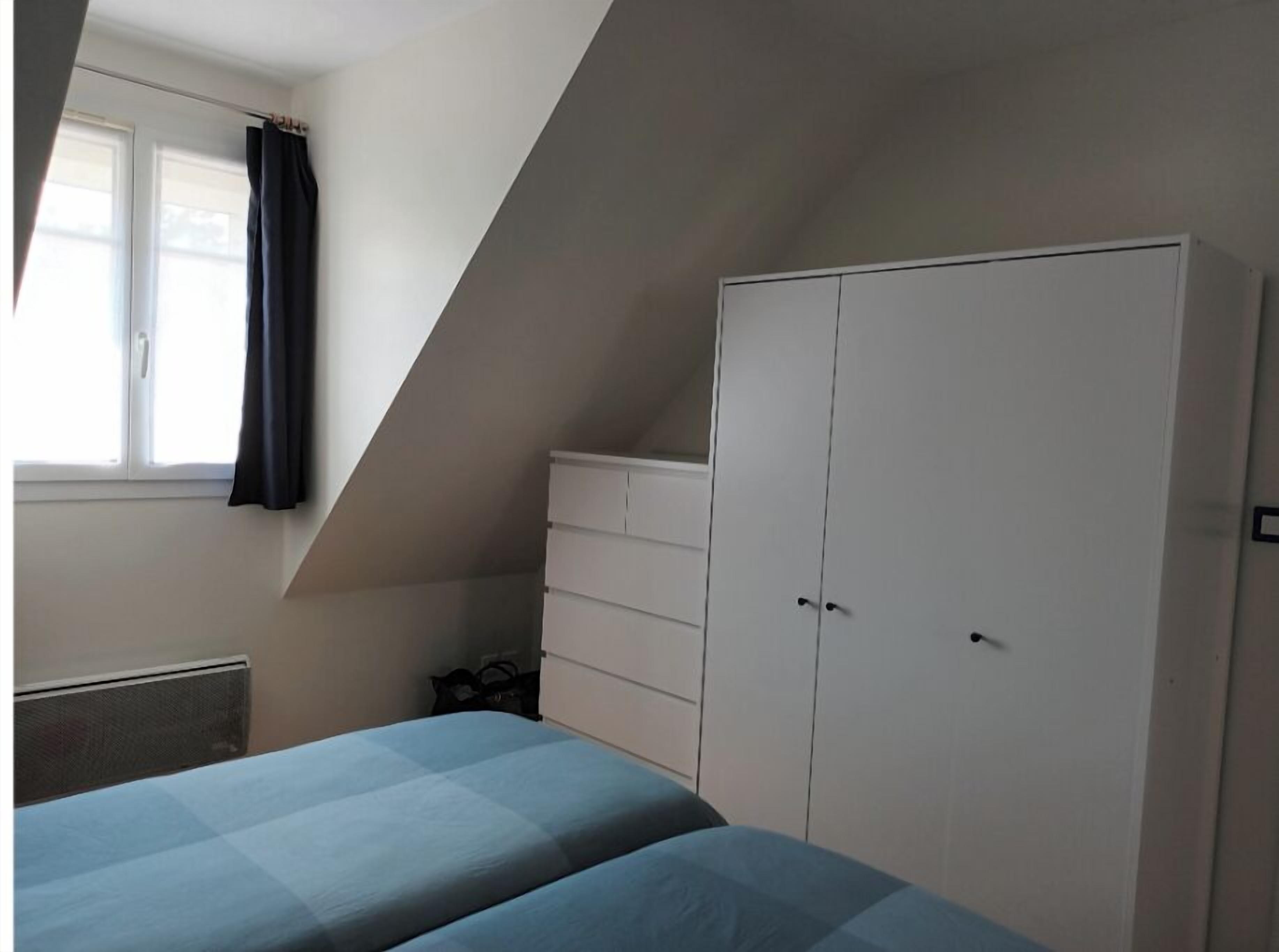 Slaapkamer met schuine kap, een bed met blauw beddengoed, een witte garderobe en een raam met een zwart gordijn.
