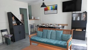 Ein modernes Wohnzimmer mit einer Couch, einem Holztisch, einem Essbereich mit Tisch und Stühlen, einer Küchenzeile mit Schränken, einem Fernseher und Kunstwerken an der Wand.