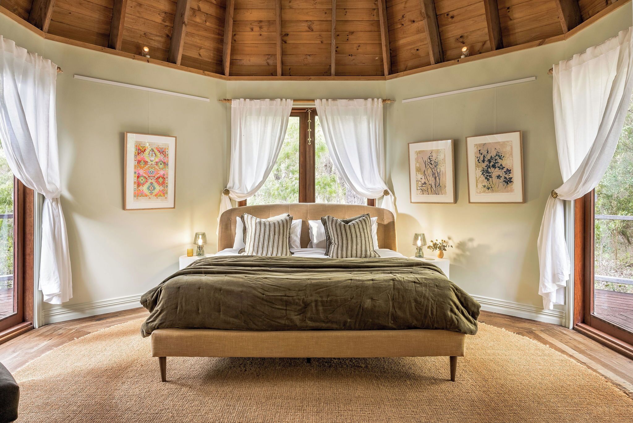 Ein Schlafzimmer mit Holzboden, einem großen Bett mit grüner Bettwäsche, weißen Vorhängen und gerahmten Kunstwerken an den Wänden.