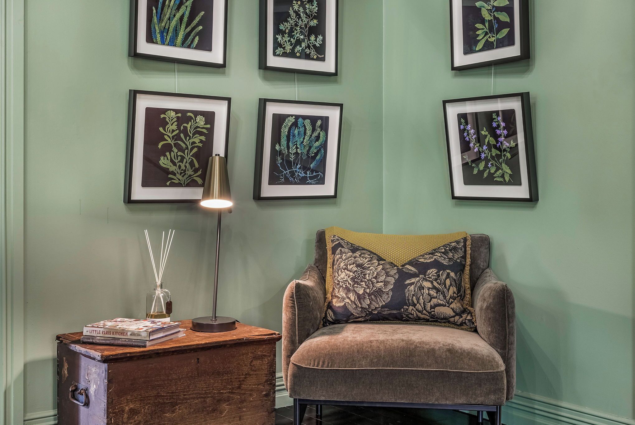 Een kamer met een groene wand, een fauteuil met een bloemenkussen, een klein houten tafeltje met een lamp en een glazen pot, en ingelijste botanische prints aan de wand.