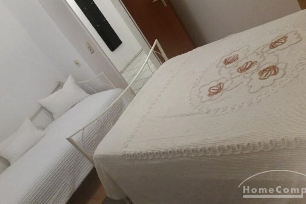 Een net opgemaakt bed met een witte dekbedovertrek en kussens, een nachtkastje en een wandlamp.