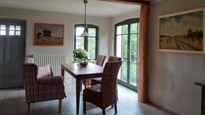 Un espace repas comprenant une table, des chaises et un luminaire suspendu. Il y a deux fenêtres avec des cadres verts et une porte.